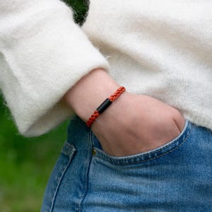 Peut inclure: Un bracelet en cordon tressé orange doté d'une perle noire cylindrique et lisse au centre. Le bracelet est porté au poignet, présentant un design simple avec une texture tissée orange vif et une pièce d'accent noire unie contrastante.