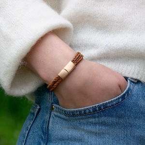 May include: A braided tan leather bracelet featuring a smooth, rectangular metallic clasp. The bracelet consists of two thin, intertwined leather cords that meet at a polished, solid-colored metal closure, creating a simple and clean design.