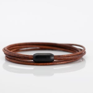 May include: A multi-strand bracelet composed of thin, brown leather cords wrapped in a circular formation. The cords are secured together by a single, smooth, matte black cylindrical bead positioned at the centre of the bracelet design.