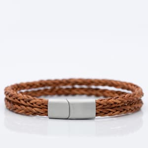 May include: A double-strand bracelet featuring two braided brown leather cords. The bracelet is secured with a smooth, rectangular, matte silver-toned metal magnetic clasp that connects the two ends of the braided leather material together.