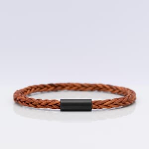 Könnte beinhalten: Ein geflochtenes braunes Lederarmband mit einem glatten, mattschwarzen zylindrischen Metallverschluss. Das Armband ist kreisförmig und weist eine durchgehend gleichmäßige geflochtene Textur auf, die durch das massive schwarze Metallteil verbunden ist.