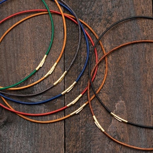 Könnte beinhalten: Ein Set aus sieben Lederkordelketten mit goldenen Verschlüssen. Die Kordeln sind in verschiedenen Farben, darunter Braun, Rot, Blau, Grau und Grün.