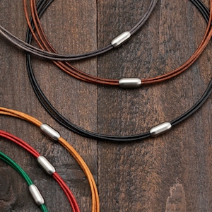 Op de afbeelding: Vier lederen koordsnoeren met zilveren magnetische sluitingen. De kettingen liggen op elkaar op een houten oppervlak. De koordsnoeren zijn zwart, bruin, beige en rood en groen.