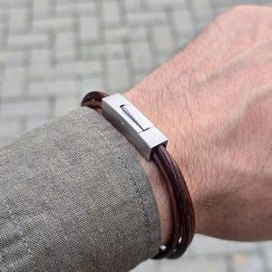 May include: A brown leather bracelet with a silver clasp. The bracelet is made of multiple strands of leather and has a simple, elegant design.