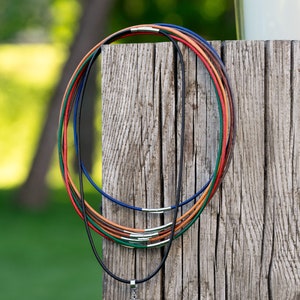 May include: A set of five leather cord necklaces in various colors, including black, blue, green, orange, and brown, are stacked on a wooden surface. The necklaces are all tied together at the top and have a silver clasp at the bottom.