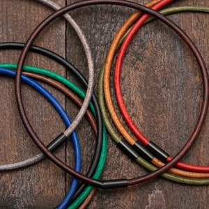 May include: A collection of leather cords in various colours, including brown, red, green, and blue, with black metal clasps. The cords are arranged in a circle on a wooden surface.