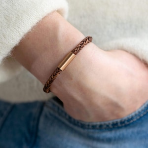 Op de afbeelding: Een bruin leren armband met een gouden metalen sluiting. De armband is gevlochten en heeft een eenvoudig ontwerp.