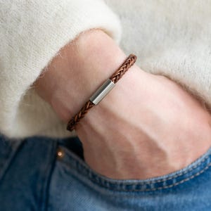 Könnte beinhalten: Ein braunes geflochtenes Lederarmband mit einem silbernen Metallakzent.