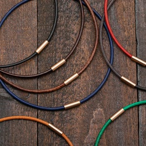 以下が含まれることがあります： 黒、茶色、青、赤、緑など、さまざまな色のミニマリストネックレスのコレクション。各ネックレスは、細い革紐と金色の金属アクセントが特徴です。ネックレスは素朴な木製の表面に配置されています。