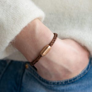 Könnte beinhalten: Ein braunes Lederarmband mit einem goldenen Metallverschluss. Das Armband ist geflochten und hat ein schlichtes Design.