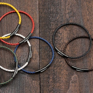 May include: Six leather bracelets in different colours, including red, yellow, white, blue, green, and brown, with black metal clasps. The bracelets are arranged on a wooden surface.
