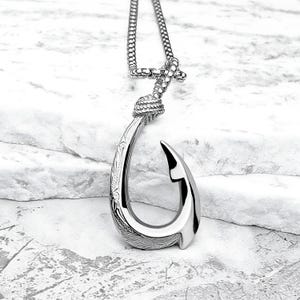 May include: A silver necklace with a detailed fish hook pendant. The hook is intricately designed with floral patterns and has a knotted chain.