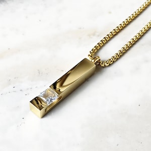 May include: A gold chain necklace with a pendant featuring a rectangular bar with a clear square-shaped gemstone set into the center.
