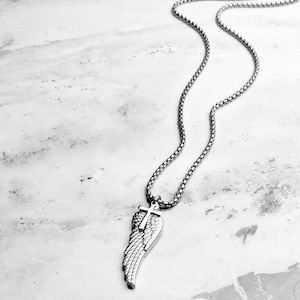May include: A silver necklace with a pendant shaped like a wing and a cross. The chain is a box link style. The wing pendant has a detailed feather design. The necklace is shown on a marble-effect surface.