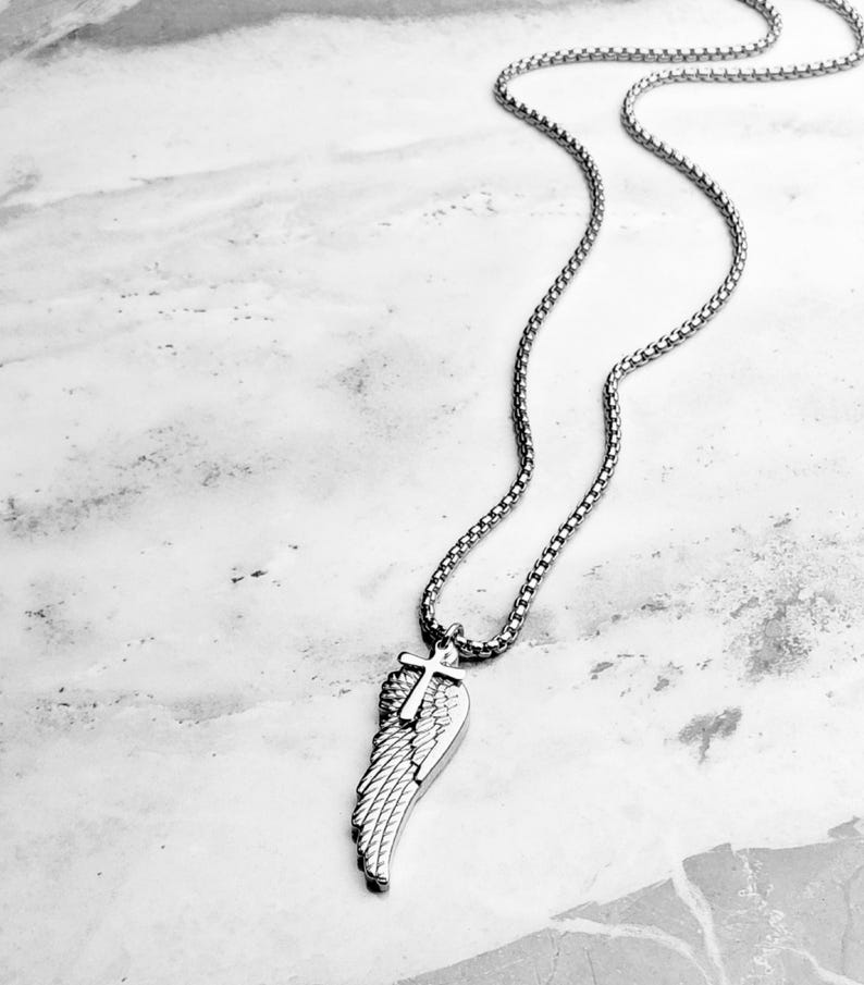 May include: A silver necklace with a pendant shaped like a wing and a cross. The chain is a box link style. The wing pendant has a detailed feather design. The necklace is shown on a marble-effect surface.