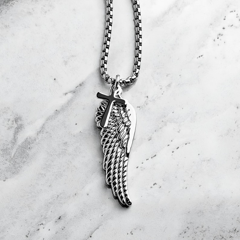 May include: A silver-coloured necklace with a pendant featuring a cross and a wing design. The chain is a box link style. The pendant has etched feather patterns. The necklace is displayed on a white and grey marble surface.
