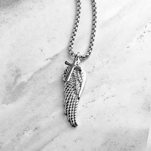 May include: Silver-coloured necklace with a pendant featuring a cross and a wing design. The pendant hangs from a box chain. The necklace is shown against a marble background.