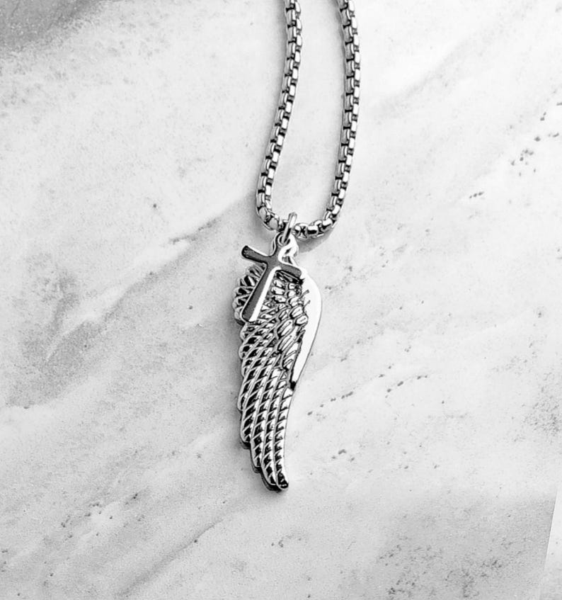 May include: Silver-coloured necklace with a pendant featuring a cross and a wing design. The pendant hangs from a box chain. The necklace is shown against a marble background.