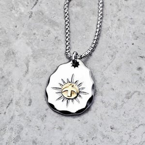 Puede incluir: Un collar de cadena de plata con un colgante de plata que presenta un sol estilizado con un pájaro dorado en su interior. El colgante tiene un aspecto texturizado y martillado.