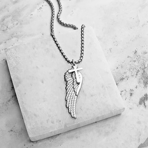 May include: A silver necklace with a pendant combining a wing and a cross. The wing is intricately detailed with feather patterns, and the cross sits above it. The chain is a linked design. The background is a light-coloured, textured surface.