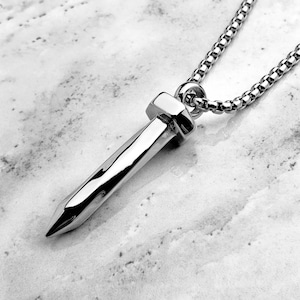 Puede incluir: Un collar de plata con un colgante de clavo de plata. El clavo es largo y puntiagudo, con una cabeza cuadrada.