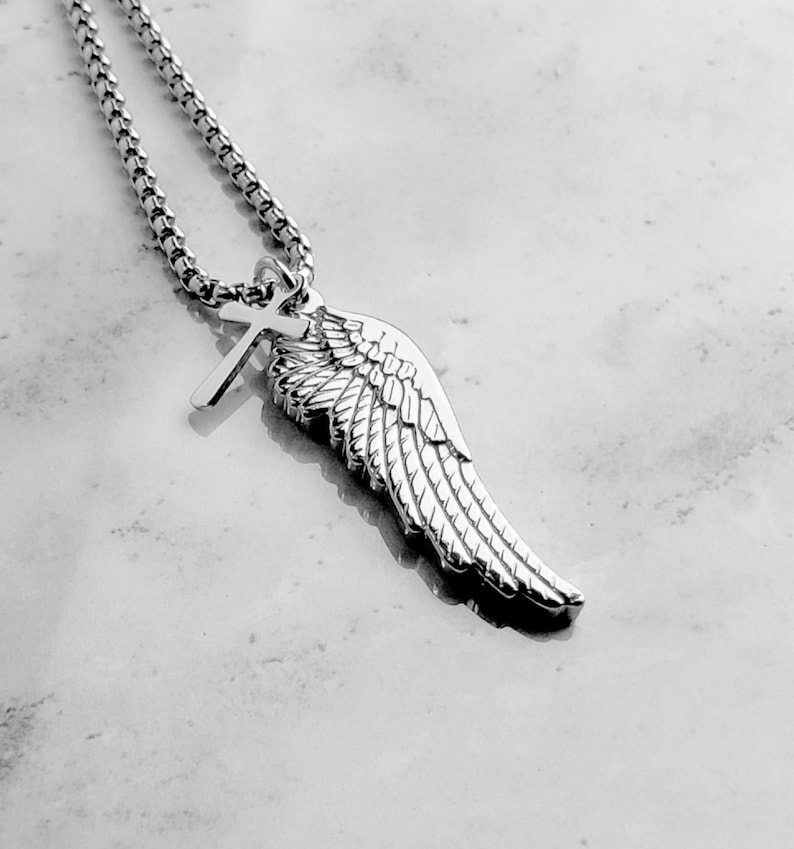 May include: A silver necklace with a pendant shaped like a wing and a cross. The wing has detailed feathering. The chain is a linked design. The pendant is highly reflective, and the necklace is set against a light-coloured background.