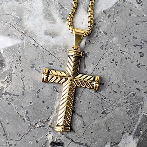 Puede incluir: Un collar con un colgante de cruz de tono dorado con un patrón de chevron detallado. La cadena es de estilo cadena de caja.