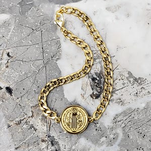 May include: Gold-colored chain bracelet with a round pendant. The pendant features an image of a religious figure and the text "S. BENEDICTI. P.S.M.V. - S.M.Q.L. IVB. - PAX". The bracelet has a lobster claw clasp.