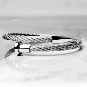 以下が含まれることがあります： ねじれたケーブルデザインのシルバーブレスレット。ブレスレットは、磨かれた金属製の留め金とエンドキャップを備えています。ケーブルはしっかりと織り込まれ、テクスチャード加工の外観を作り出しています。ブレスレットは反射面の上に表示されています。