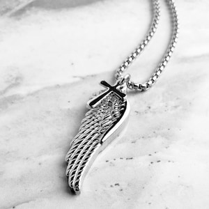 May include: A silver pendant necklace with a wing and cross design. The wing has detailed feathering, and the cross sits at the top. The chain is a linked style. The background is a marble-effect surface.