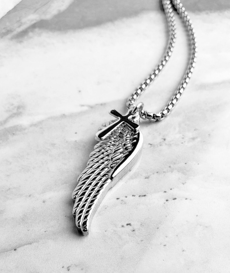 May include: A silver pendant necklace with a wing and cross design. The wing has detailed feathering, and the cross sits at the top. The chain is a linked style. The background is a marble-effect surface.