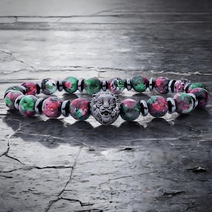May include: A beaded bracelet featuring a silver lion head charm. The bracelet is made of round beads in shades of red, green, and black. The lion head charm is centered on the bracelet. The bracelet is displayed on a textured, gray surface.