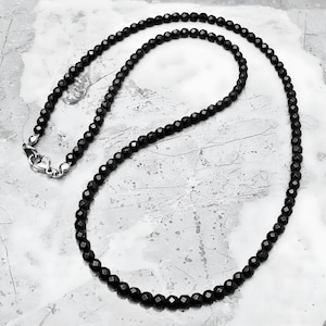 May include: A black beaded necklace with a silver clasp. The necklace is composed of small, faceted black beads, creating a sparkling effect. The necklace is displayed on a textured, light gray surface.