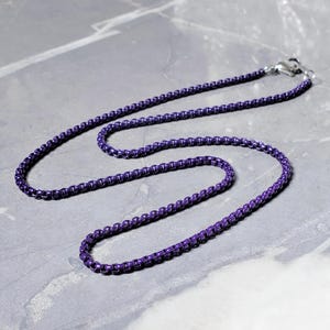 May include: A purple necklace with a box chain design. The necklace is made of metal and has a lobster clasp closure. The chain is a deep, rich purple color and is displayed on a gray surface.