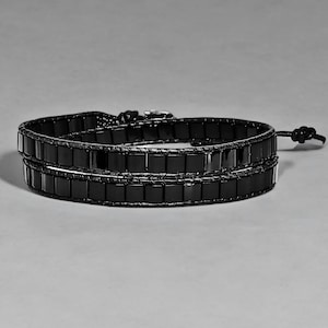 Op de afbeelding: Een zwarte wikkelarmband met rechthoekige, glanzende zwarte kralen. De armband bestaat uit twee rijen kralen, verbonden door een dunne, donkere band. Hij wikkelt twee keer om de pols en heeft een geknoopte sluiting.