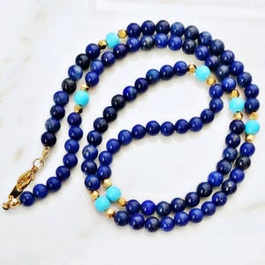 May include: A necklace featuring dark blue lapis lazuli beads, interspersed with turquoise and gold-colored accents. The necklace has a gold-colored clasp and is displayed on a white surface. The beads are approximately 8mm in diameter.