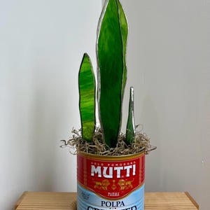 May include: A decorative stained glass plant sculpture featuring three green leaf-shaped elements with silver edging, set in a MUTTI crushed tomatoes can. The can is red and blue with text.