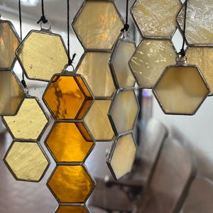May include: Honeycomb-shaped stained glass ornaments in shades of yellow and amber hang from black cords. The glass has a textured appearance, and the metal edging adds a decorative touch. The ornaments are suspended in front of a blurred background.