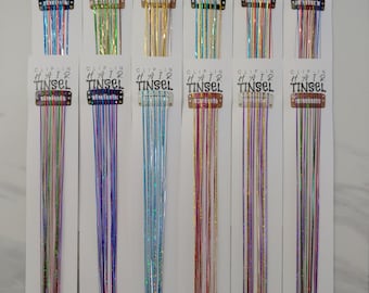 May include: A selection of clip-in hair tinsel in a range of colours, such as blue, green, gold, and purple. Each strand of tinsel is attached to a small clip. The packaging reads "CLIP-IN HAIR TINSEL".