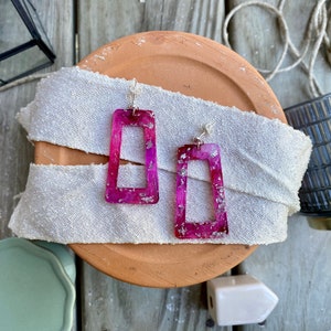 Op de afbeelding: Een paar rechthoekige hangende oorbellen met een basis van doorschijnende magenta hars met ingebedde zilverkleurige metaalschilfers. Elke oorbel hangt aan een kleine zilverkleurige metalen steker met een bijpassende sluiting.