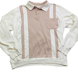 May include: A long-sleeved polo shirt in cream, tan, and white. The shirt features a cream-colored collar and sleeves, with a tan center panel and a small pocket. White vertical stripes run down the front.
