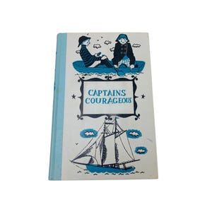 May include: Vintage hardcover book titled "Captains Courageous." The cover features illustrations of two figures in a boat, a sailboat, and the title in a decorative frame. The book has a light blue spine and a white cover with blue and black illustrations.