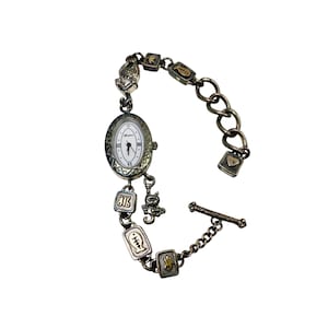 May include: Silver-toned watch bracelet with an oval watch face and decorative charms. The watch face has a white background with black hands and Roman numerals. Charms include a cat, fish, and heart. The bracelet has a toggle clasp.