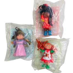 May include: Three vintage Cabbage Patch Kids dolls in their original plastic packaging. One doll wears a red dress and blue boots, another a purple dress, and the third a red and white dress with a teddy bear apron. Each doll has a unique hairstyle.