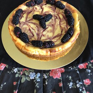 May include: A swirl cheesecake with a creamy white and purple filling topped with fresh blackberries. The cheesecake is on a gold plate.