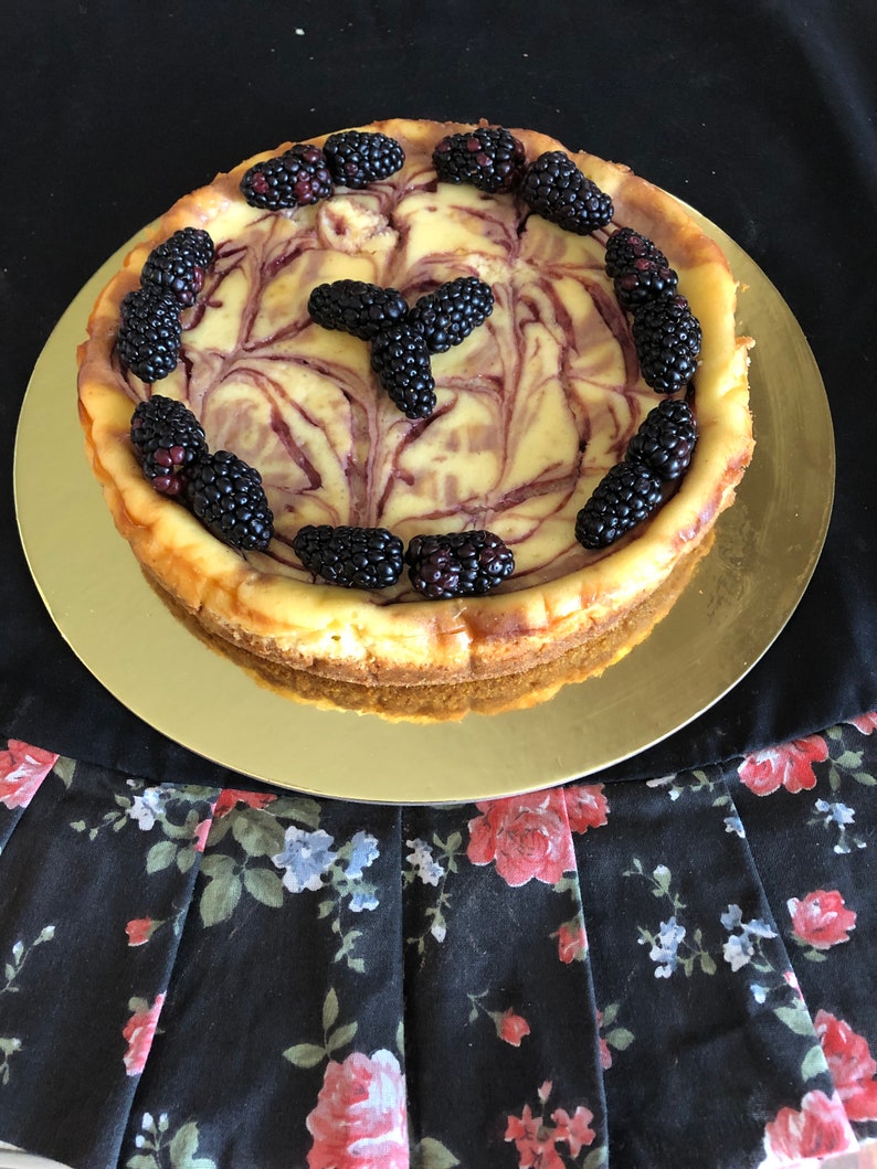 May include: A swirl cheesecake with a creamy white and purple filling topped with fresh blackberries. The cheesecake is on a gold plate.