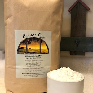 May include: A brown paper bag with a white label that reads "Rise and Shine 100% Organic Non-GMO Hard Red Wheat Flour" and includes the company's name, address, and contact information. A white ceramic bowl filled with flour sits in front of the bag.