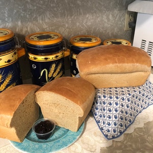 May include: Two loaves of freshly baked bread, one sliced open on a blue plate with a small dish of jam. The bread is on a white lace tablecloth. There are four blue and yellow ceramic canisters in the background.