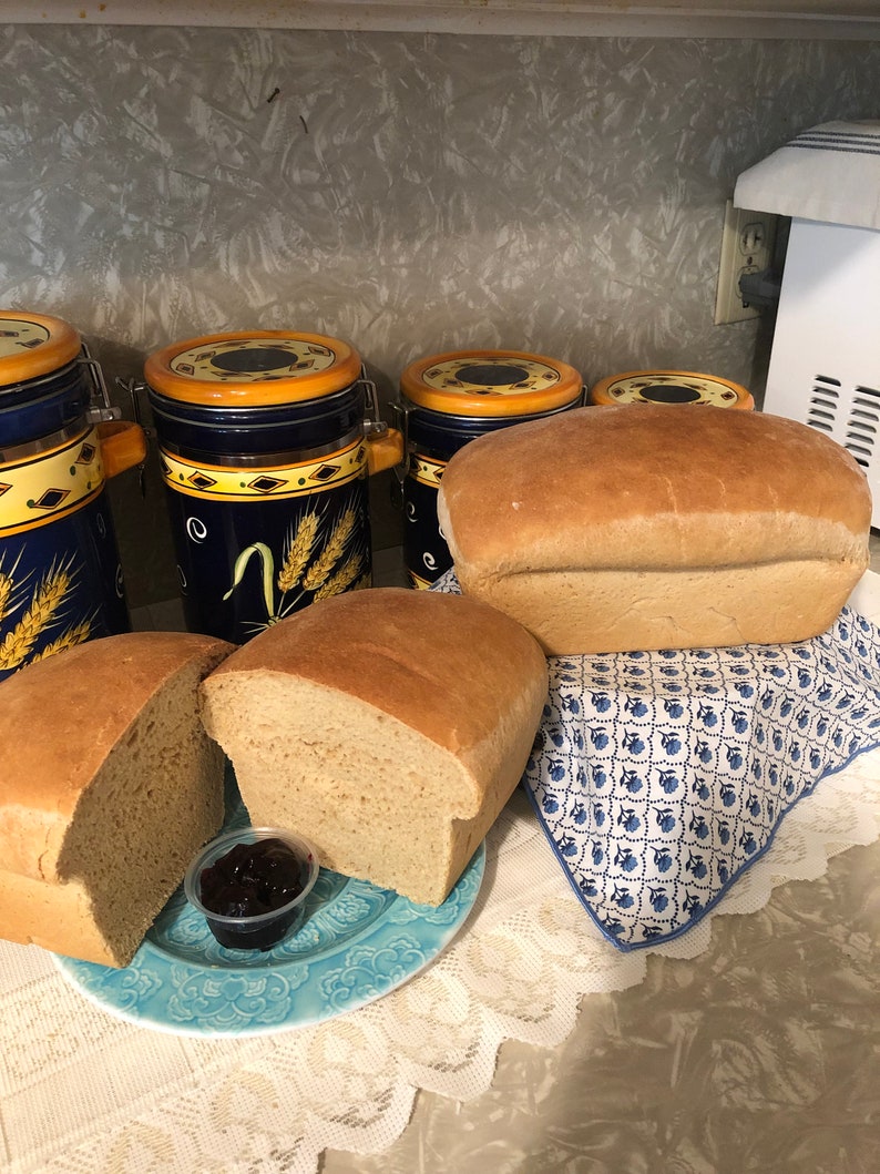 May include: Two loaves of freshly baked bread, one sliced open on a blue plate with a small dish of jam. The bread is on a white lace tablecloth. There are four blue and yellow ceramic canisters in the background.