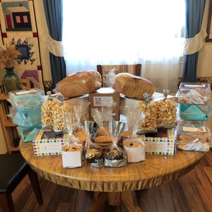 May include: A table filled with baked goods, including loaves of bread, popcorn, cookies, and brownies. The table is covered in a brown tablecloth and has a variety of containers and bags holding the food.